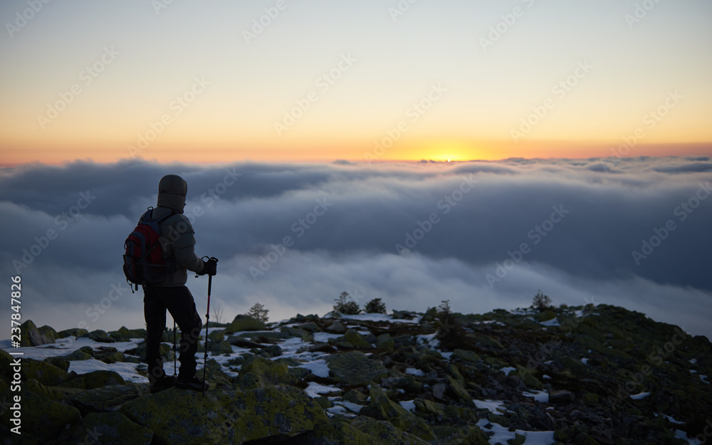 Back view of tourist in warm jacket with backpack and hiking sticks on rocky mountain peak on background of valley covered with white clouds stretching to horizon and bright morning sky at down.