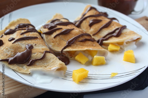 Healthy vegan wheat crepes decorated with chocolate. Wheat crepes with fresh mango and yogurt on plate and on wooden and white background. Healthy breakfast. Horizontal image. 