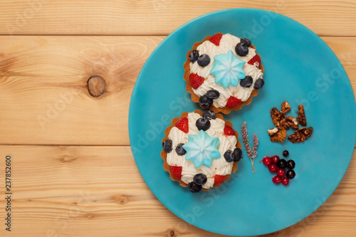Two tartlets with white butter cream, blue marshmallow on top and strawberries and blueberries on blue plate and with currant, walnuts and mint flower. The plate is on a wooden surface