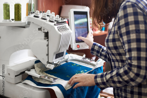 Fotografie Close-up of woman working on modern computerized specially engineered embroidery machine with multi-needle fixed embroidery head creating green floral pattern on bright blue textile detail