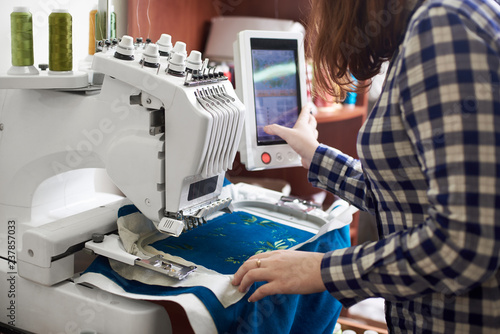 Close-up of woman working on modern computerized specially engineered embroidery machine with multi-needle fixed embroidery head creating green floral pattern on bright blue textile detail.