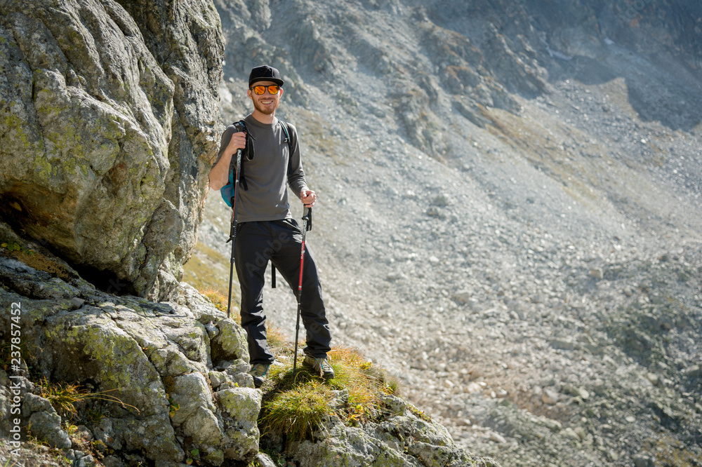 Porter of a stylish hipster traveler with a beard and a backpack in sunglasses and a cap with trekking poles stands on a rock against the backdrop of a mountain circus