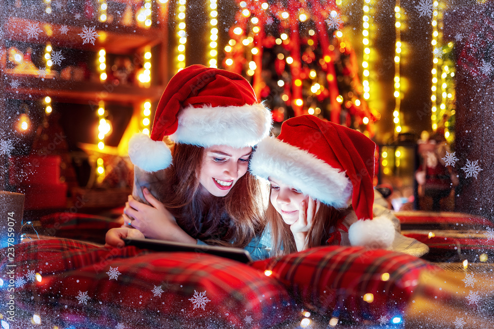 Christmas time. Smiling family mother and daughter in santas hats and pajamas watching funny video or choosing gifts on digital tablet while lie on the bed