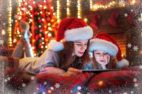 Christmas time. Smiling family mother and daughter in santas hats and pajamas watching funny video or choosing gifts on digital tablet while lie on the bed © zamuruev