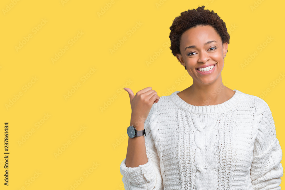 Beautiful young african american woman wearing sweater over isolated background smiling with happy face looking and pointing to the side with thumb up. Beautiful young african american woman wearing sweater over isolated background smiling with happy face looking and pointing to the side with thumb up.