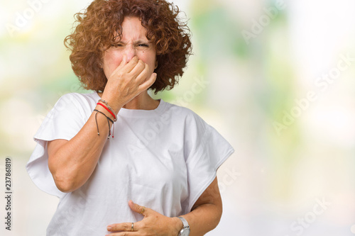 Beautiful middle ager senior woman wearing white t-shirt over isolated background smelling something stinky and disgusting, intolerable smell, holding breath with fingers on nose. Bad smells concept.