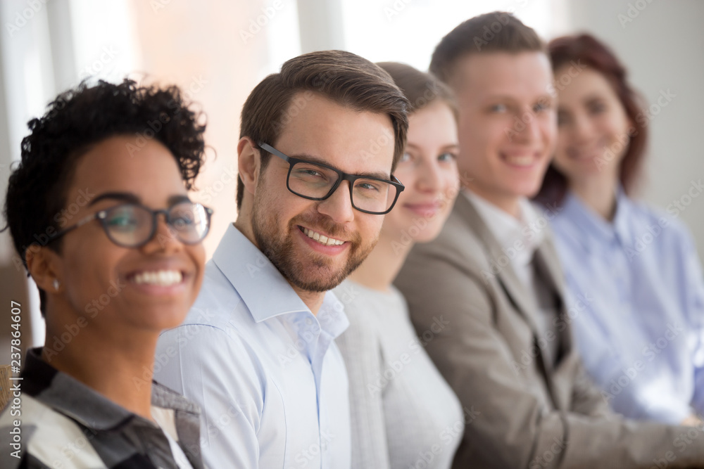 Team of multiracial millennial employees in row look at camera smiling, focus on young male worker wearing glasses posing at company briefing, portrait of diverse workers sit together at meeting Team of multiracial millennial employees in row look at camera smiling, focus on young male worker wearing glasses posing at company briefing, portrait of diverse workers sit together at meeting