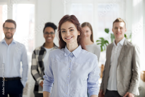 Portrait of smiling millennial female employee or worker looking at camera, happy successful woman standing foreground posing with colleagues at background, student or intern make picture in office photo
