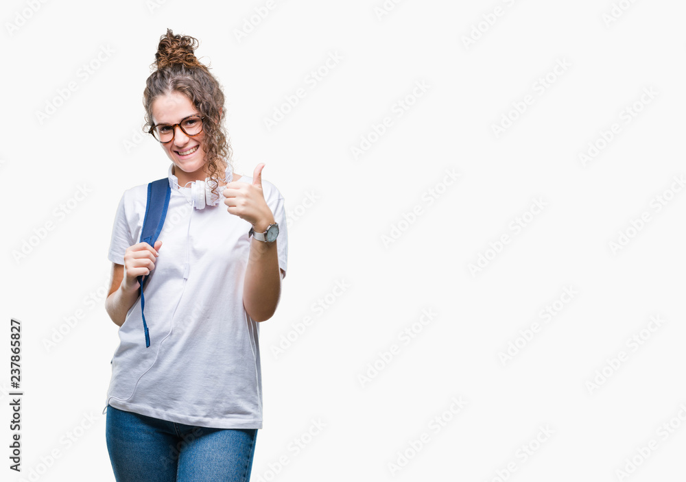 Young brunette student girl wearing backpack and headphones over isolated background doing happy thumbs up gesture with hand. Approving expression looking at the camera with showing success. Young brunette student girl wearing backpack and headphones over isolated background doing happy thumbs up gesture with hand. Approving expression looking at the camera with showing success.