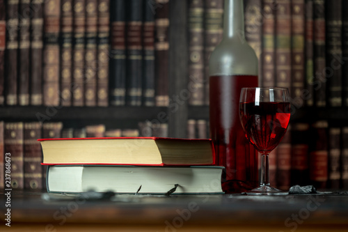 in a room full of books, on the table stands a glass and a bottle of red wine, next to them lies two books