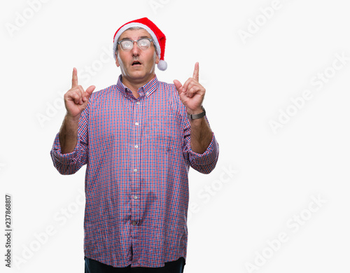 Handsome senior man wearing christmas hat over isolated background amazed and surprised looking up and pointing with fingers and raised arms.