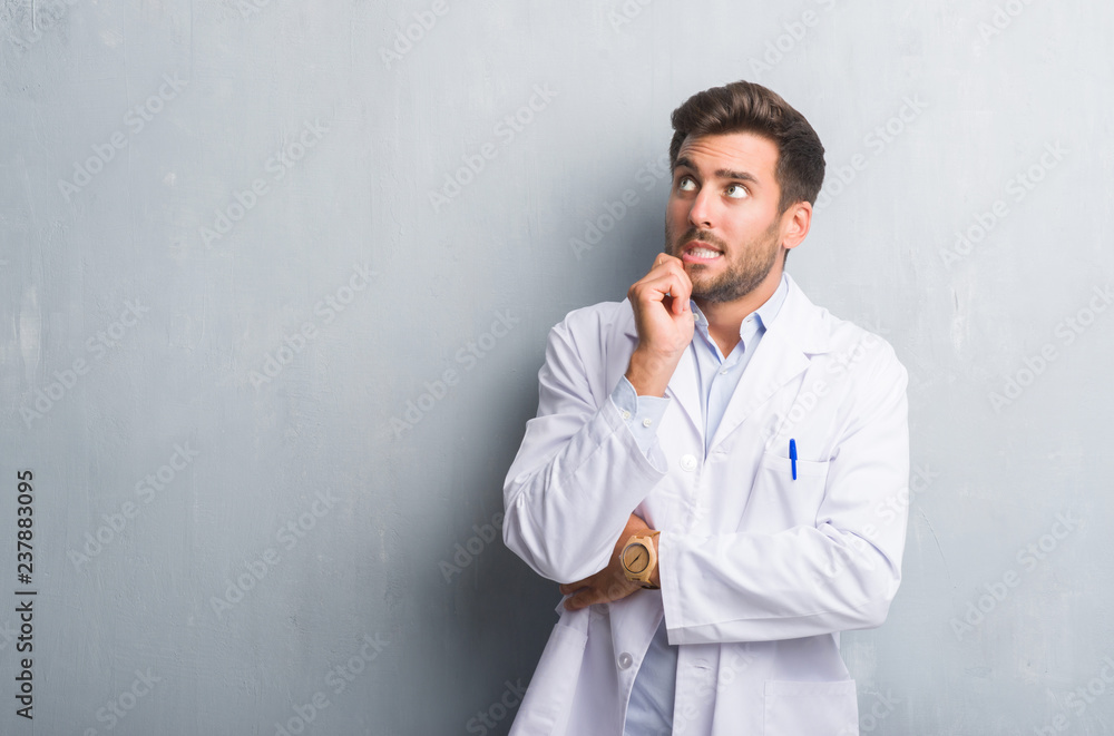 Handsome young professional man over grey grunge wall wearing white coat with hand on chin thinking about question, pensive expression. Smiling with thoughtful face. Doubt concept. Handsome young professional man over grey grunge wall wearing white coat with hand on chin thinking about question, pensive expression. Smiling with thoughtful face. Doubt concept.
