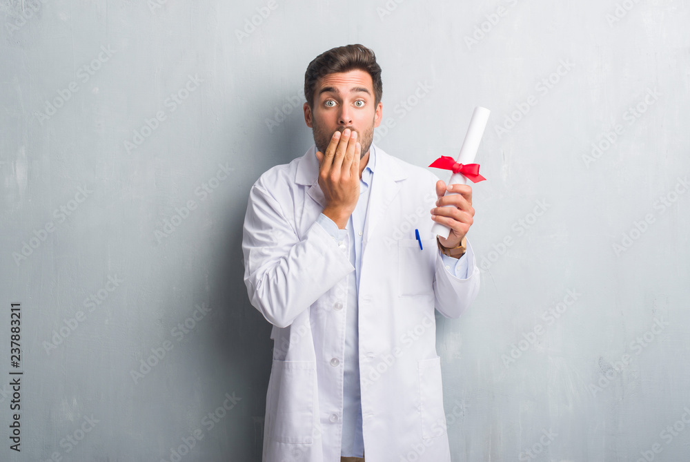 Handsome young doctor man over grey grunge wall holding diploma cover mouth with hand shocked with shame for mistake, expression of fear, scared in silence, secret concept Handsome young doctor man over grey grunge wall holding diploma cover mouth with hand shocked with shame for mistake, expression of fear, scared in silence, secret concept