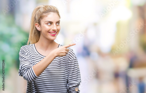 Young beautiful blonde woman wearing stripes sweater over isolated background cheerful with a smile of face pointing with hand and finger up to the side with happy and natural expression on face