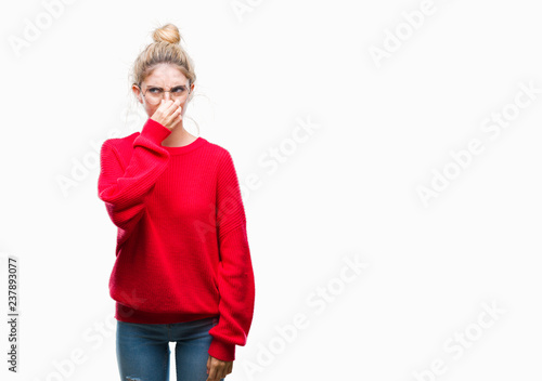 Young beautiful blonde woman wearing red sweater and glasses over isolated background smelling something stinky and disgusting, intolerable smell, holding breath with fingers on nose