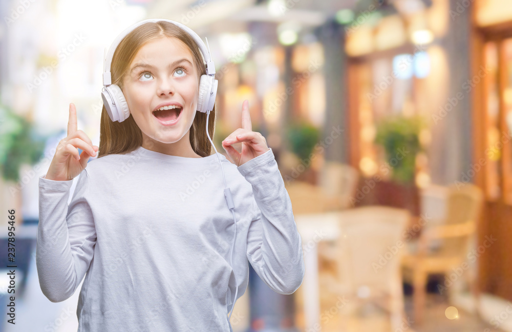 Young beautiful girl wearing headphones listening to music over isolated background amazed and surprised looking up and pointing with fingers and raised arms.