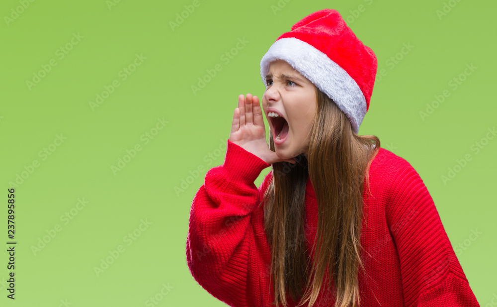 Young beautiful girl wearing christmas hat over isolated background shouting and screaming loud to side with hand on mouth. Communication concept. Young beautiful girl wearing christmas hat over isolated background shouting and screaming loud to side with hand on mouth. Communication concept.