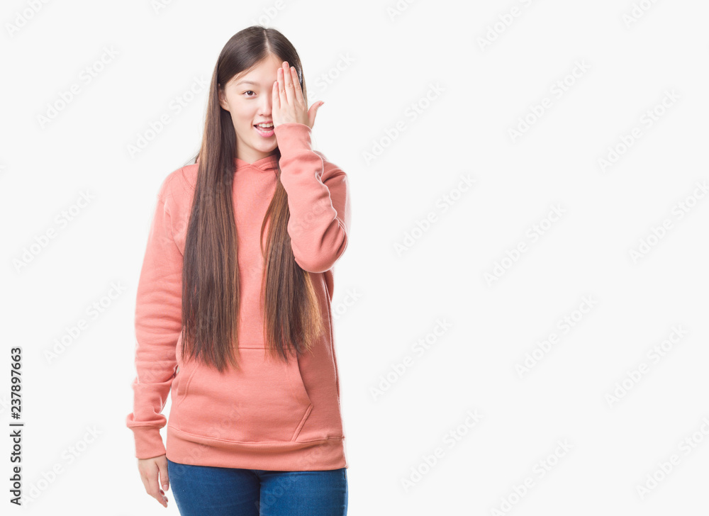 Young Chinese woman over isolated background wearing sport sweathshirt covering one eye with hand with confident smile on face and surprise emotion. Young Chinese woman over isolated background wearing sport sweathshirt covering one eye with hand with confident smile on face and surprise emotion.