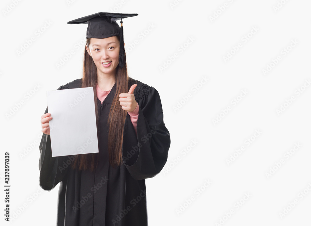 Young Chinese woman wearing graduate uniform holding paper degree happy with big smile doing ok sign, thumb up with fingers, excellent sign Young Chinese woman wearing graduate uniform holding paper degree happy with big smile doing ok sign, thumb up with fingers, excellent sign