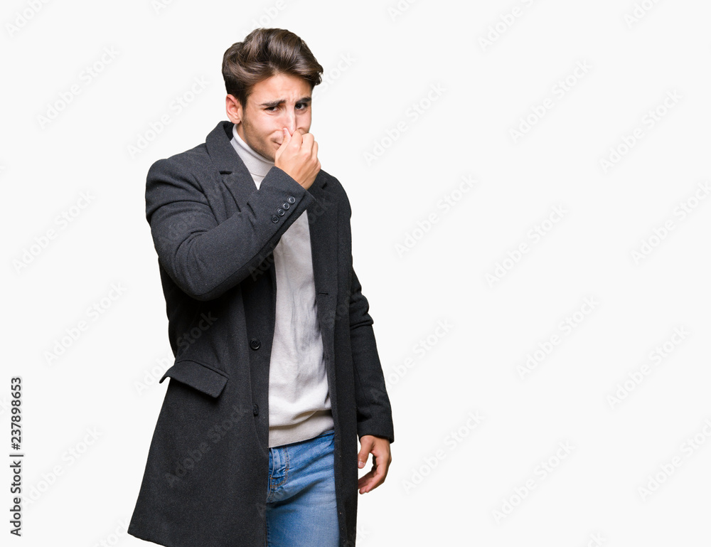 Young elegant man wearing winter coat over isolated background smelling something stinky and disgusting, intolerable smell, holding breath with fingers on nose. Bad smells concept.
