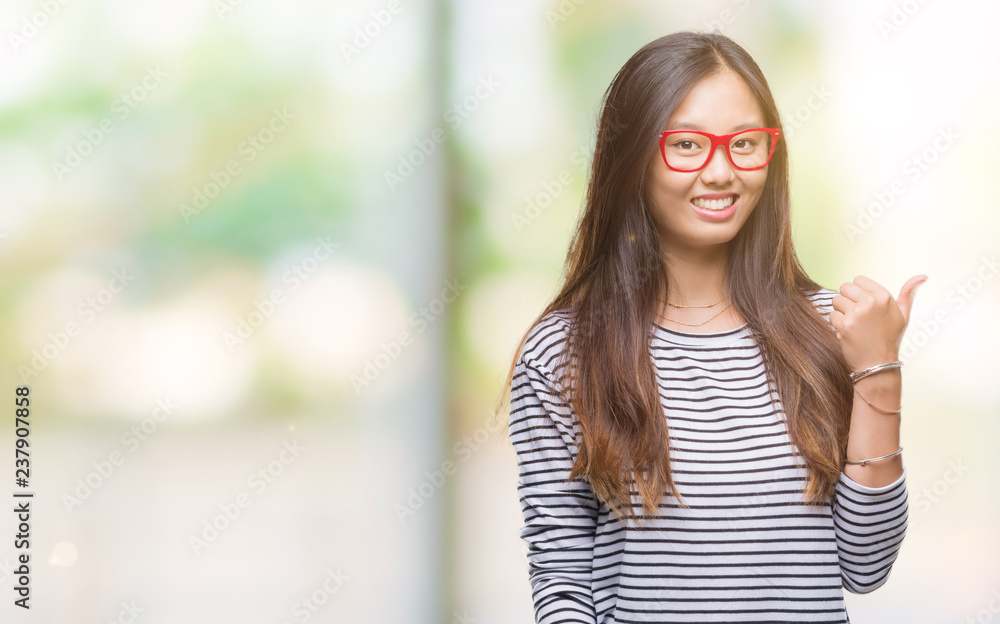 Young asian woman wearing glasses over isolated background smiling with happy face looking and pointing to the side with thumb up.