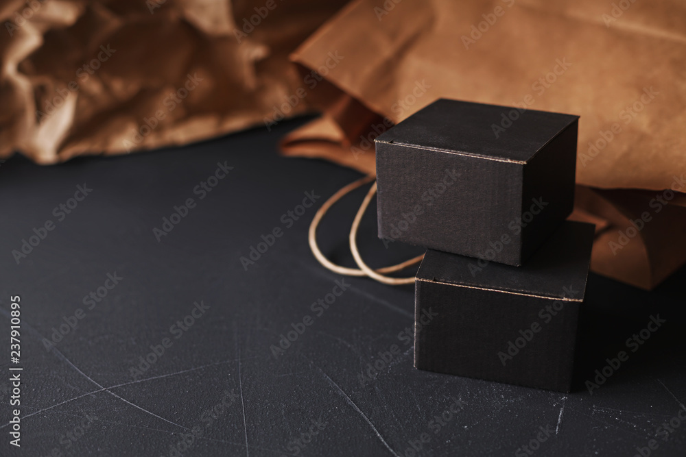 Two matte black carton gift boxes laying on a black painted scratched surface, craft paper bag blurred on background