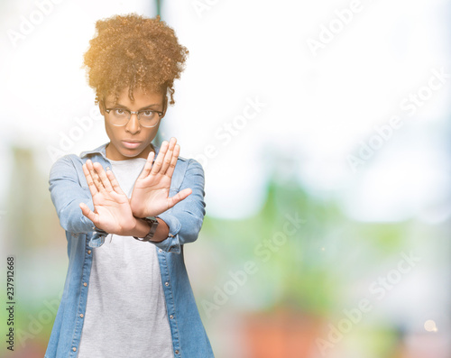Beautiful young african american woman wearing glasses over isolated background Rejection expression crossing arms and palms doing negative sign, angry face © Krakenimages.com