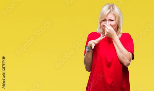 Young beautiful blonde woman wearing red t-shirt over isolated background smelling something stinky and disgusting, intolerable smell, holding breath with fingers on nose. Bad smells concept.