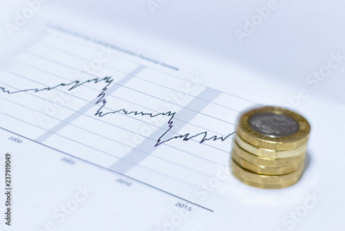 GBP British Currency Pound Coins spread out and stacked on top of 2d graph data sheet showing exponential growth over time