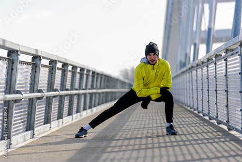 Young athlete man stretching his muscles before running on bridge on sunny winter day