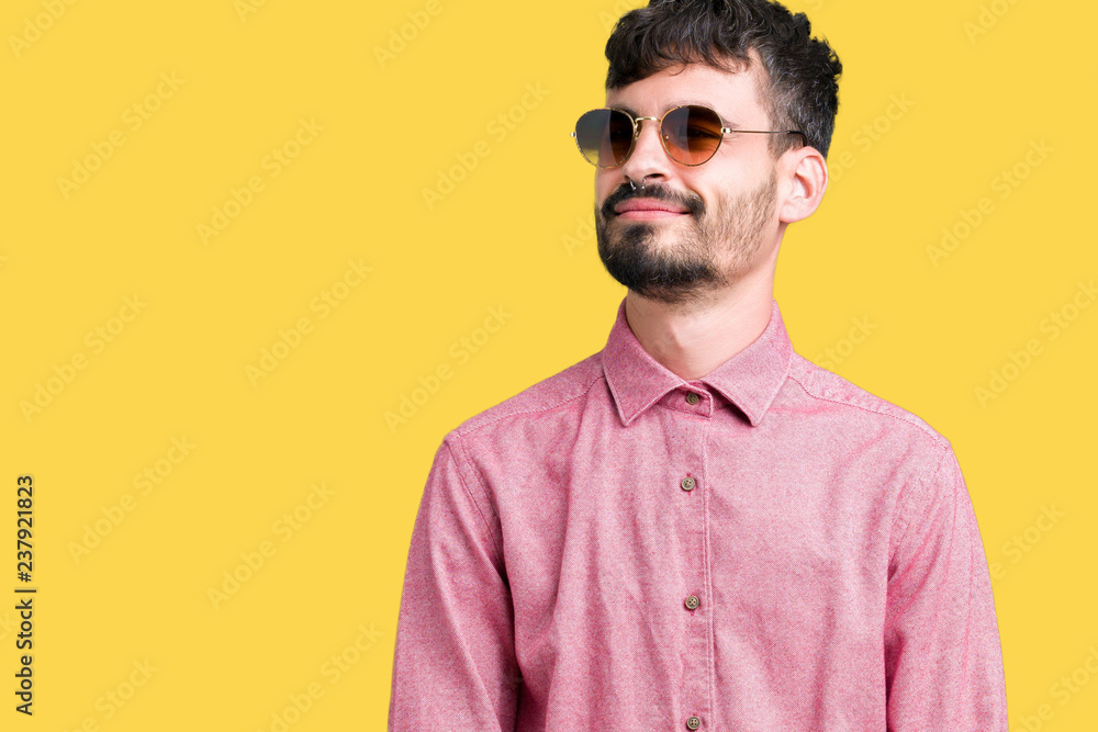 Fototapeta premium Young handsome man wearing sunglasses over isolated background looking away to side with smile on face, natural expression. Laughing confident.