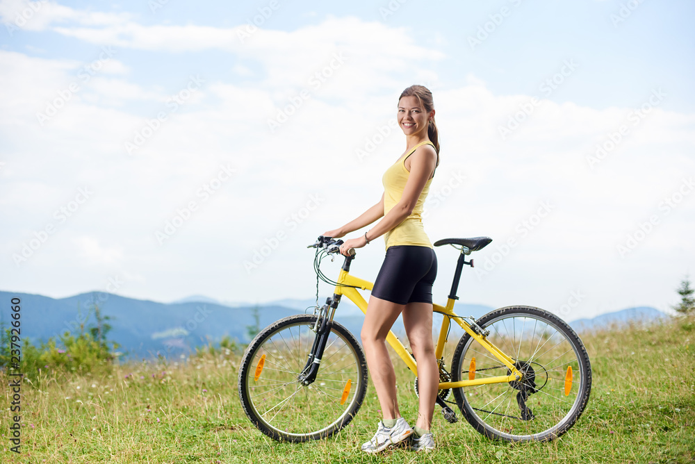 Attractive happy girl biker standing on a grassy hill with yellow mountain bicycle, enjoying summer day in the mountains. Outdoor sport activity, lifestyle concept