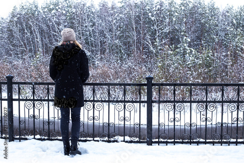 a girl in a black coat stands with her back alone on the waterfront near the forest in winter