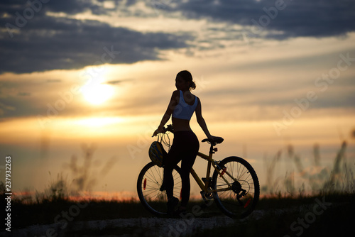 Slender female cyclist standing near bicycle and looking at sun and marvelous landscapes. Silhouette of sporty woman posing on trail in grass. Concept of motivation and healthy lifestyle.
