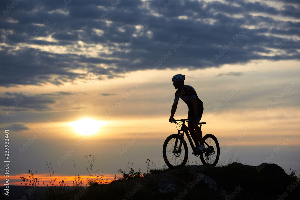 Fototapeta premium Silhouette of energetic and active cyclist standing on bicycle. Athletic man in helmet and sportswear posing on hill in twilight. Amazing background of sunset and cloudy evening sky.