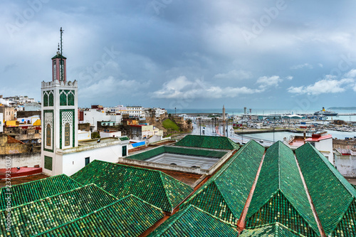 TANGIER / MOROCCO - NOVEMBER 2018: tiled roofs of buildings one of the many mosques of the medina of Tangier with a minaret and the kasbah / medina old quarter behind during the rain.