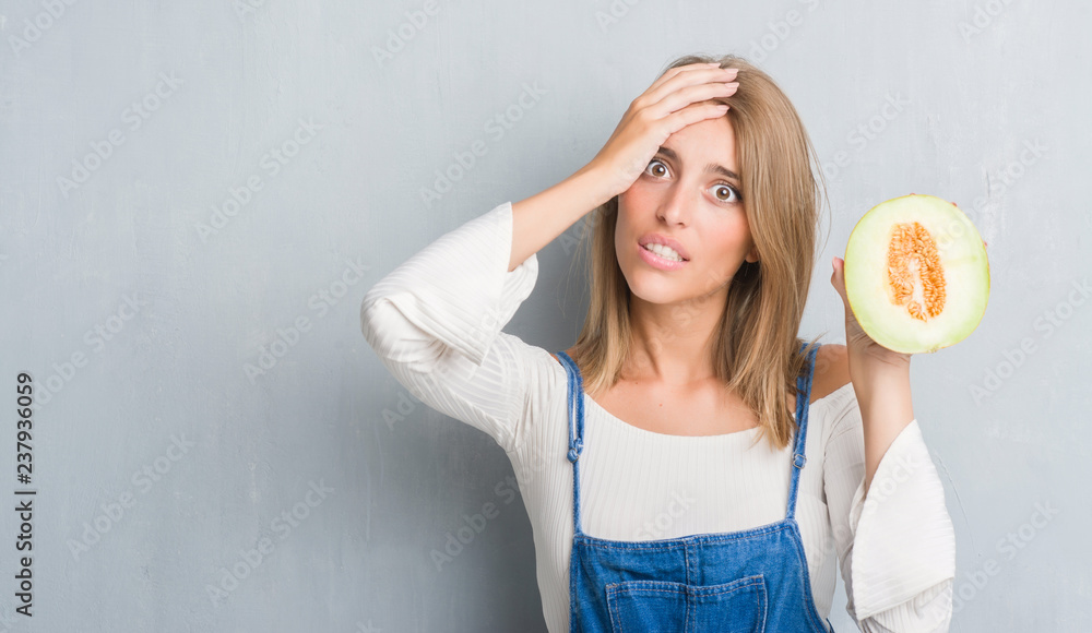 Beautiful young woman over grunge grey wall holding fresh cantaloupe melon stressed with hand on head, shocked with shame and surprise face, angry and frustrated. Fear and upset for mistake.