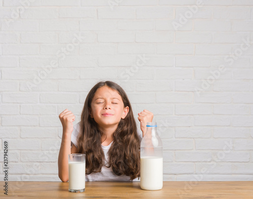 Young hispanic kid sitting on the table drinking a glass of milk screaming proud and celebrating victory and success very excited, cheering emotion
