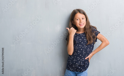 Young hispanic kid over grunge grey wall smiling with happy face looking and pointing to the side with thumb up. © Krakenimages.com