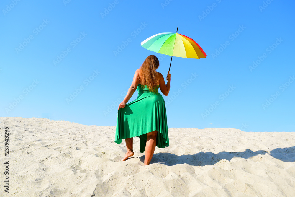 Rear view of a fat woman in a green dress with a colorful umbrella in the desert against the sky.