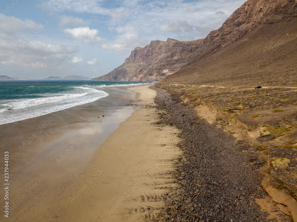 Vista aerea della spiaggia di Famara, Lanzarote, isole Canarie, Spagna. Risco di Famara, rilievo, montagne a picco sull’Oceano Atlantico. Strada non asfaltata che costeggia la costa