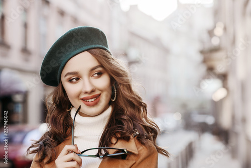 Outdoor close up portrait of young beautiful happy smiling woman with long hair wearing stylish autumn clothes, green beret,  model walking in street of european city. Copy, empty space for text