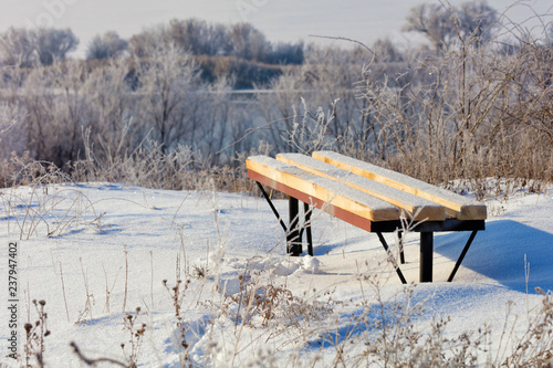 A wooden bench sprinkled with snow stands against the backdrop of the river bank.