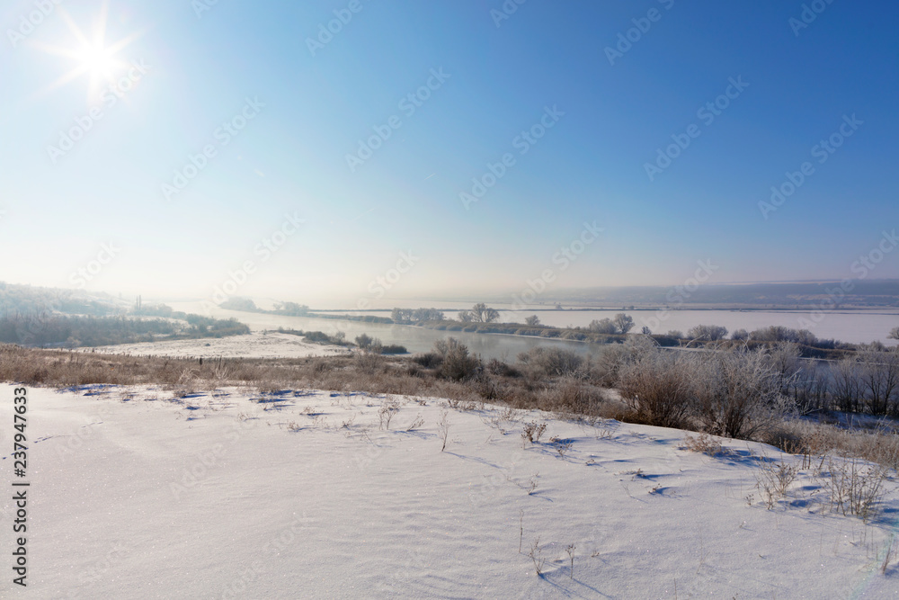Bright sun with a haze in the blue sky illuminates the river and snow-covered fields in the frosty morning