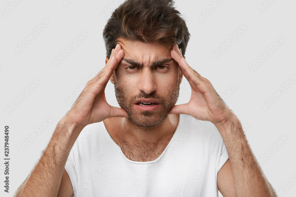Exasparated unshaven man keeps hands on temples, has terrible headache, feels tired of constant work, has dark stubble, wears casual t shirt, isolated over white studio background. Close up shot. Exasparated unshaven man keeps hands on temples, has terrible headache, feels tired of constant work, has dark stubble, wears casual t shirt, isolated over white studio background. Close up shot.