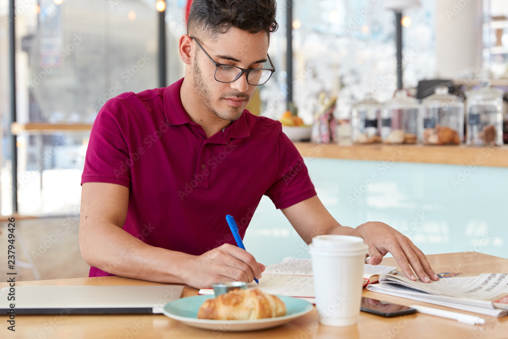 Young businessman rules company remotely, writes information in notepad. Handsome man wears optical glasses, studies university timetable, rewrites something from newspaper, models in bistro Young businessman rules company remotely, writes information in notepad. Handsome man wears optical glasses, studies university timetable, rewrites something from newspaper, models in bistro