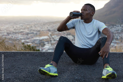 Fototapeta Naklejka Na Ścianę i Meble -  African American man drinks fresh water from bottle, rests on asphalt, sits against mountain background outdoor, feels relaxed, dressed in casual t shirt, sneakes and trousers. Relaxation concept