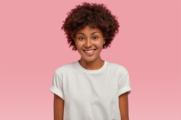 Indoor shot of attractive young African American woman with toothy tender smile, shows white teeth, dressed in casual mock up t shirt, expresses positive emotions, stands against pink background
