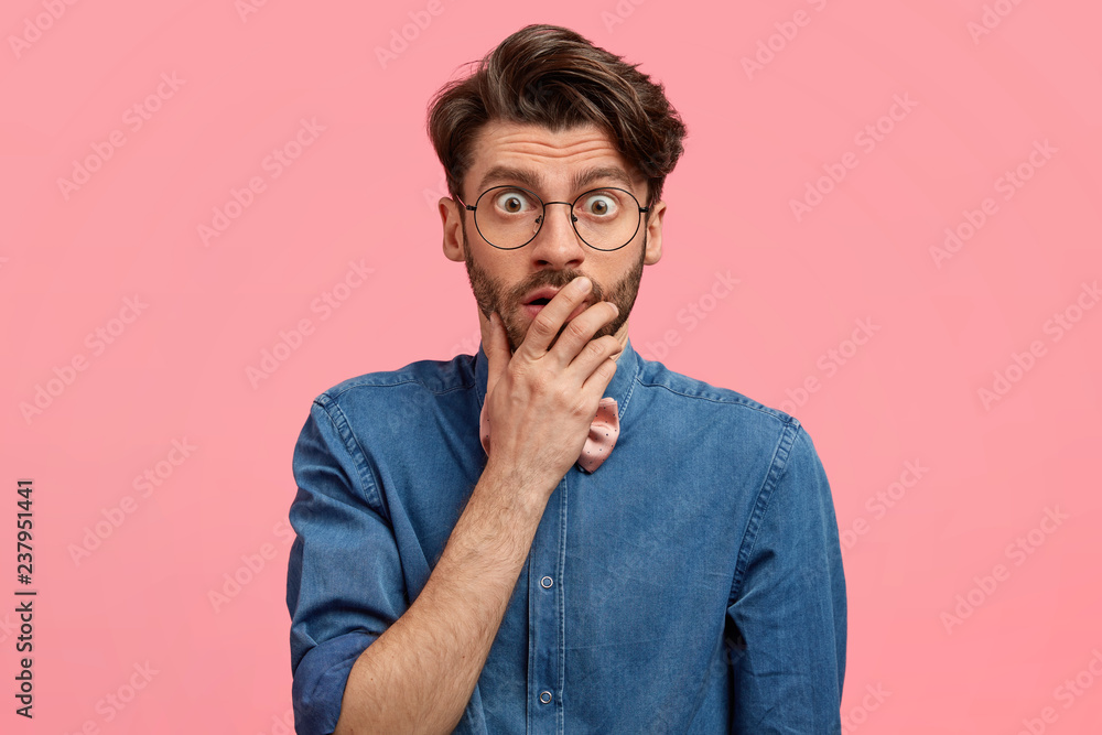 Shocked handsome young man covers mouth, has eyes popped out, stares through spectacles, wears denim shirt, models over pink background. People, unexpected reaction and surprisement concept. Shocked handsome young man covers mouth, has eyes popped out, stares through spectacles, wears denim shirt, models over pink background. People, unexpected reaction and surprisement concept.