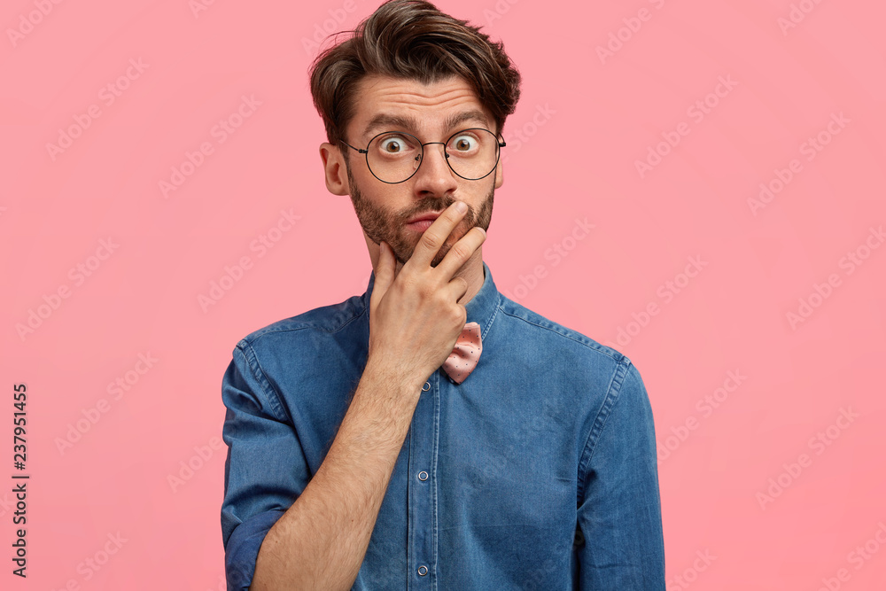 Photo of surprised frightened Caucasian bearded man keeps hand on mouth, looks with eyes full of shock, wears stylish denim shirt, notices something unbelievable, models against pink studio wall Photo of surprised frightened Caucasian bearded man keeps hand on mouth, looks with eyes full of shock, wears stylish denim shirt, notices something unbelievable, models against pink studio wall